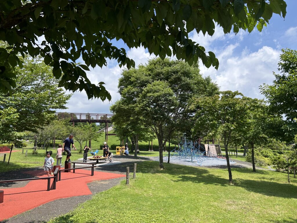桜公園の遊具エリアで遊ぶ子どもたちの風景。緑に囲まれた広々としたスペースに、滑り台やクライミングネットが見える。