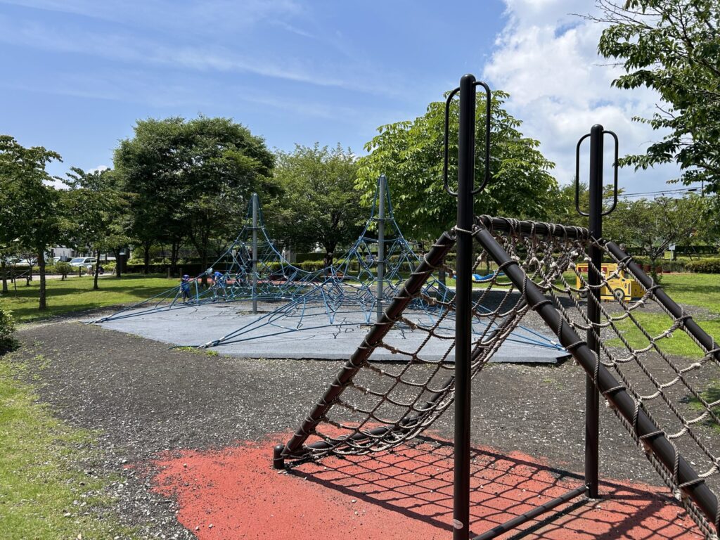 公園の遊具エリアで、青空の下にあるクライミングネットと遊び場の風景。緑に囲まれた自然の中で子どもたちが遊んでいる様子。
